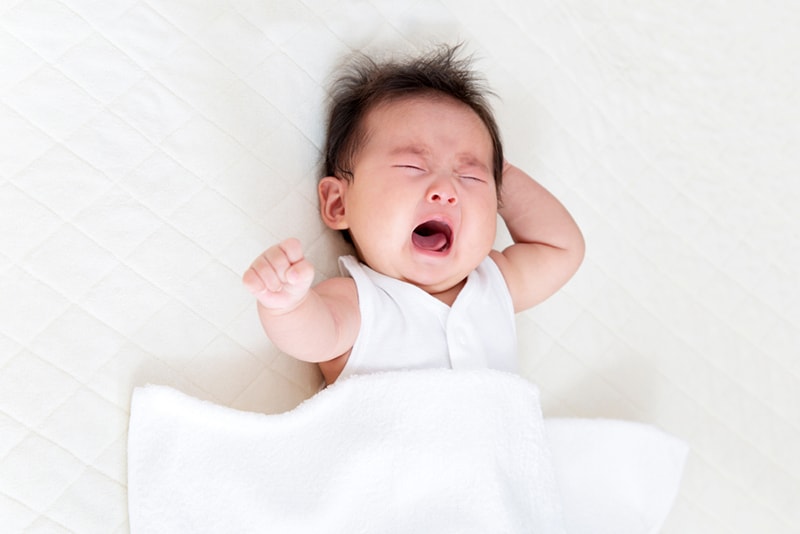 baby with long hair crying while lying on bed covered with white sheet