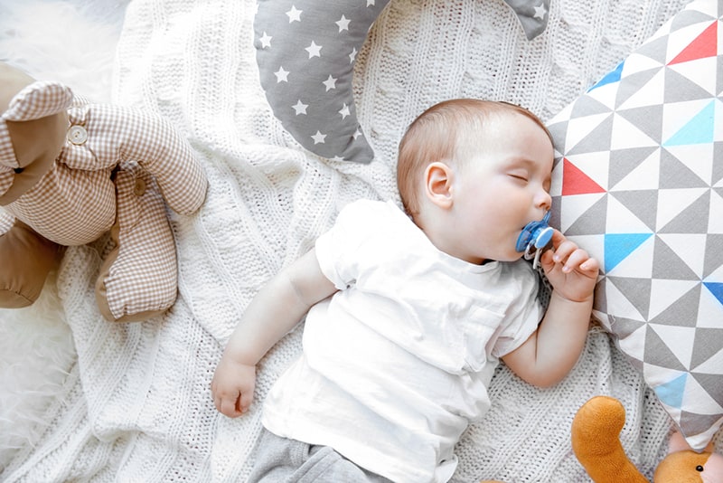 cute baby boy with pacifier sleeping on blanket with pillows and toys