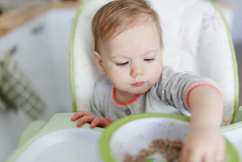 cute baby girl sitting in the high chair and eating food with hands