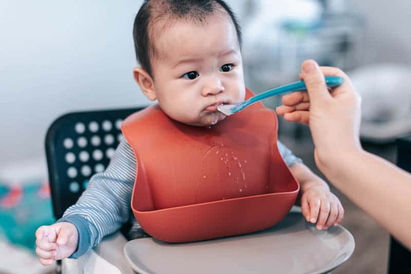 cute baby wearing red bib while getting fed by mother