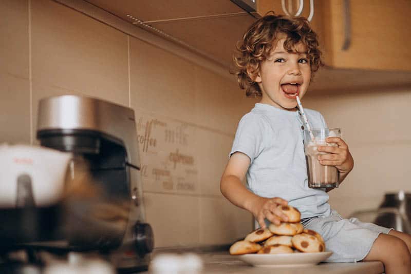 cute little boy sitting on the kitchen with plate of cookies and a chocolate milk