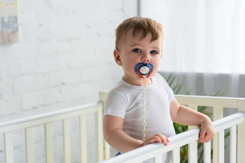 cute little boy with pacifier in mouth standing in the crib