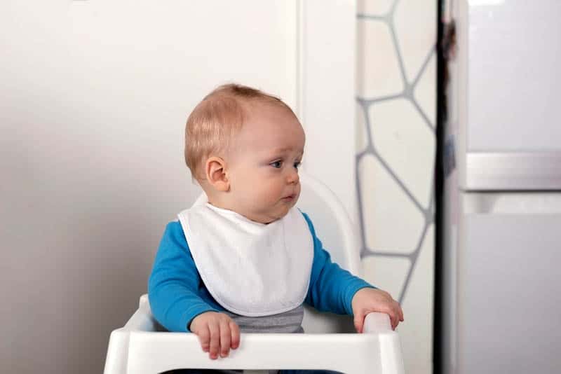 cute sad baby boy sitting in a high chair wearing bib and waiting for food