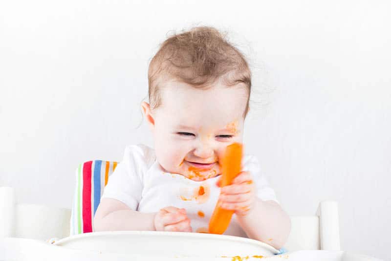 funny little baby holding a carrot in hand and smiling covered with food