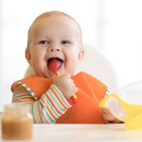 cute happy baby sitting in a high chair and eating food with a spoon