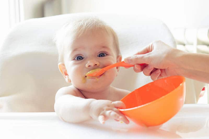mother feeding baby with food from orange bowl and orange spoon