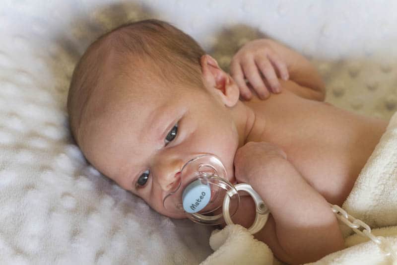 naked newborn baby sucking pacifier and lying on bed covered with cozy blanket