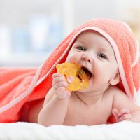 cute baby covered with orange towel chewing teether on bed