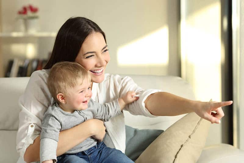 smiling mother sitting on the couch with baby boy in her lap and pointing finger at distance