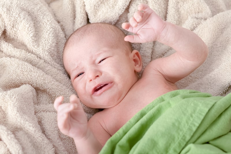 upset newborn baby crying in bed covered with green blanket