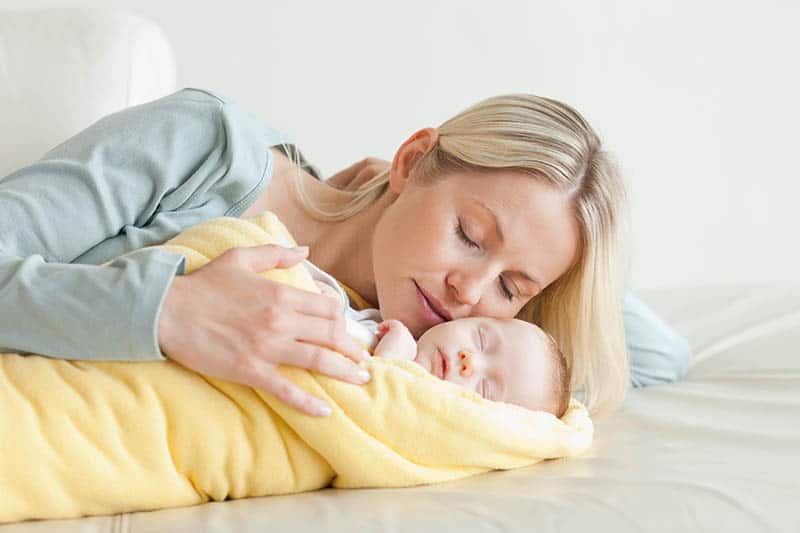 woman gently kissing her sleeping baby wrapped into yellow blanket on the bed