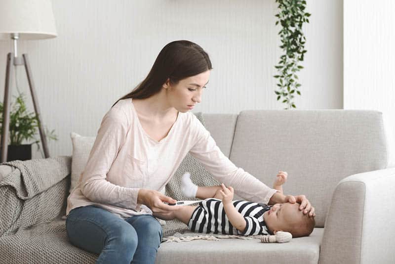 worried mother sitting with baby on the couch and checking temperature of baby with thermometer