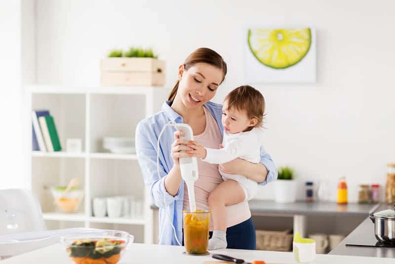 young mother holding baby girl and blending food in the kitchen