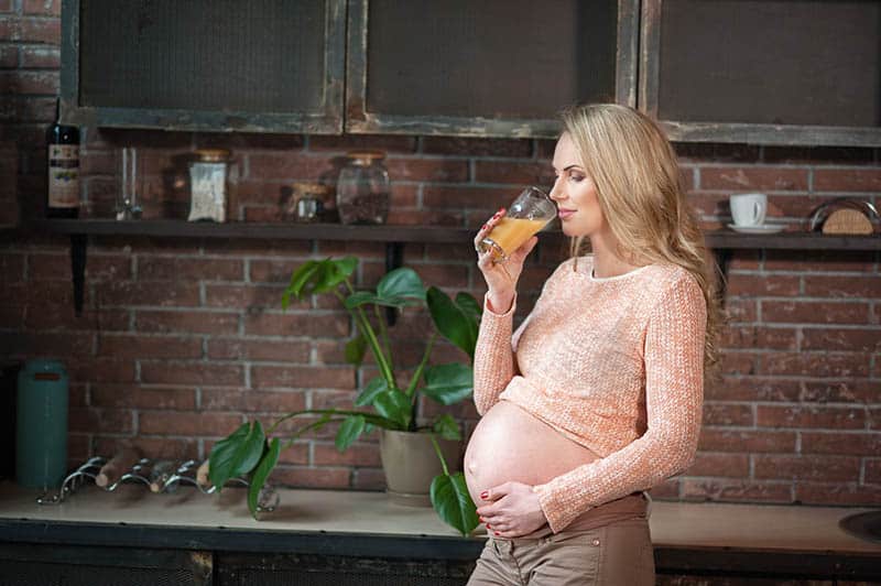 young pregnant woman standing in the kitchen and drinking orange shake