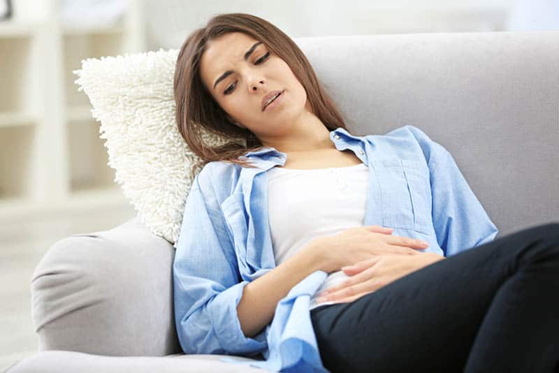 young woman feeling abdominal pain and sitting on the couch at home