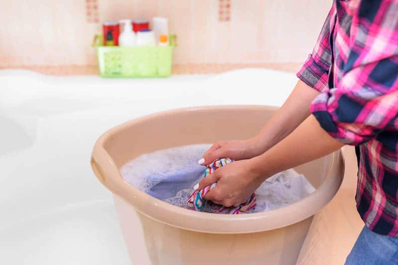 young woman in shirt handwashing baby clothes in gallon bucket in bathtub
