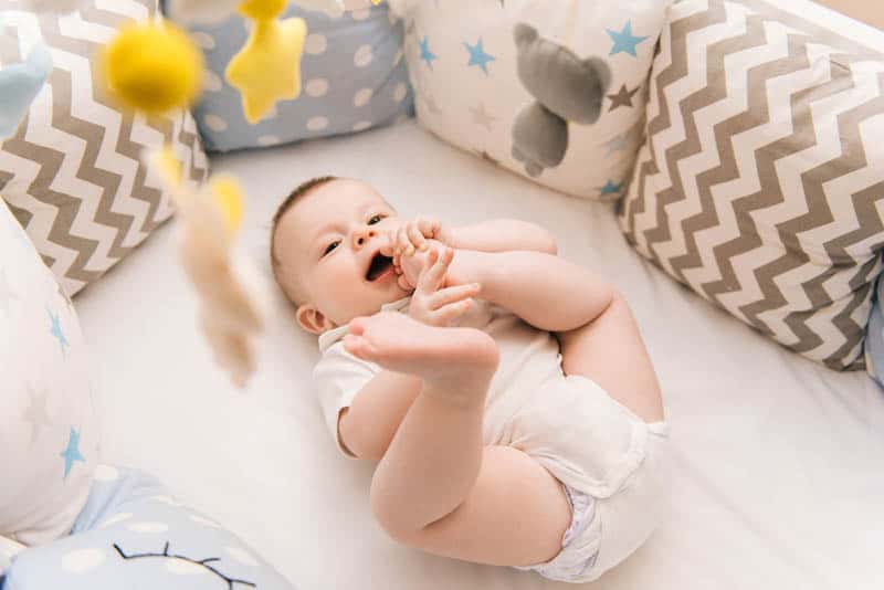 baby lying in crib with pillows around and chewing his feet