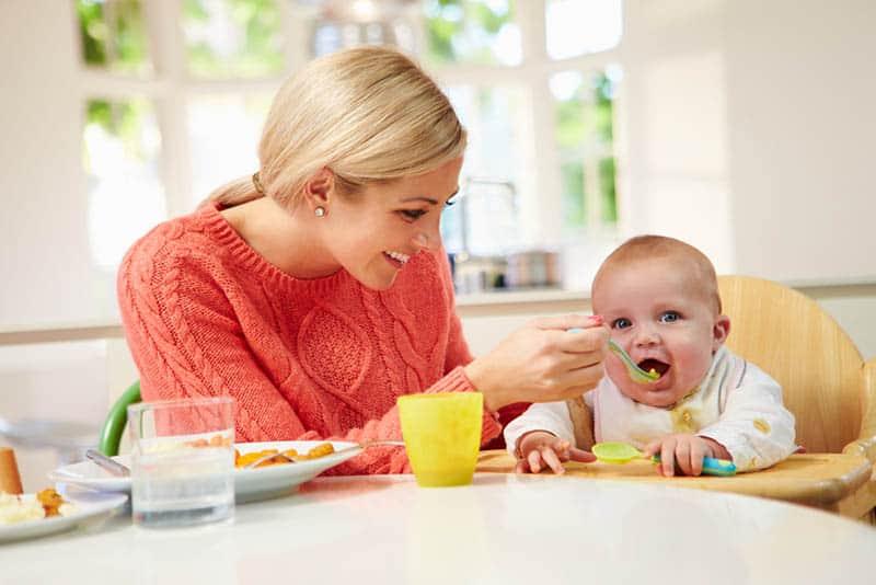 beautiful young mother feeding baby in high chair in the dining room
