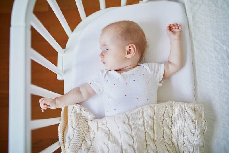 cute baby sleeping in a bassinet with white frame covered with cozy blanket