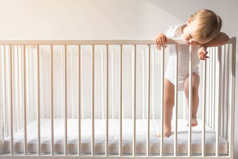 cute baby wearing white onesie standing in the crib and biting a rail