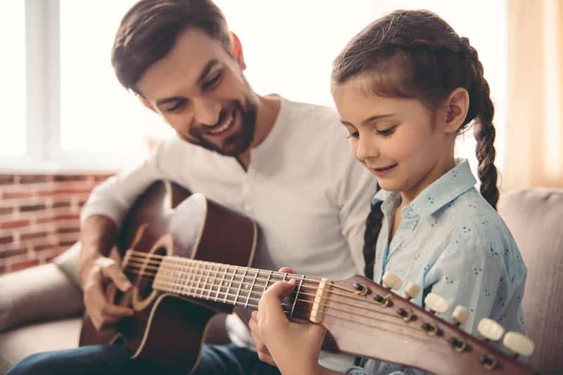 father teaching daughter to play a guitar at home on the couch