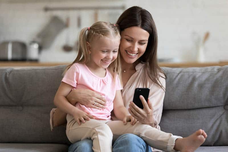 happy mother with cute daughter on the couch holding mobile phone