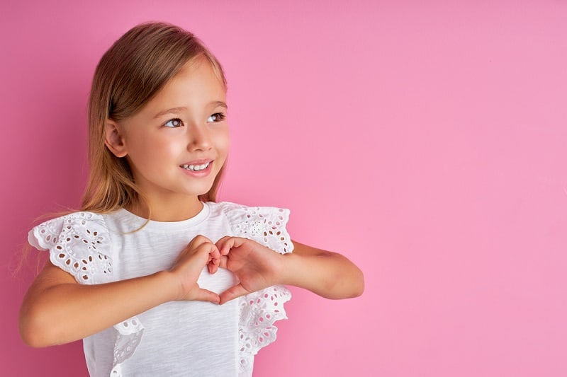 little girl making a heart shape with her hands while posing in front of a pink background