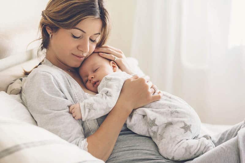 pretty mother holding sleeping baby on her chest on the bed