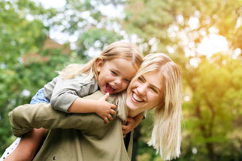 smiling blond mother carrying her daughter on back outdoor in the park