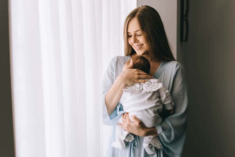 smiling young mother carrying newborn baby at home by the window