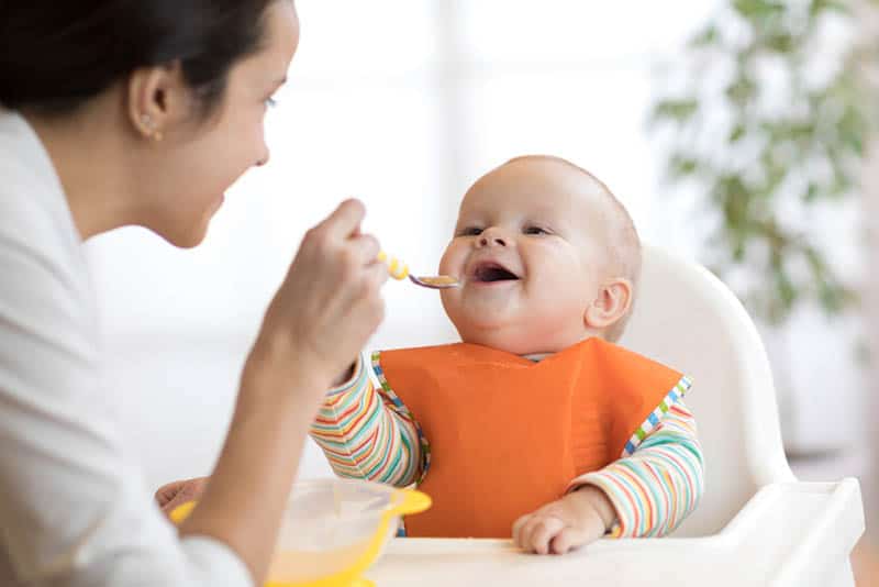 young mother feeding her smiling baby in high chair