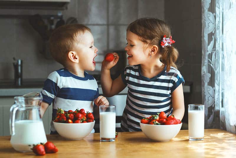 cute little girl feeding her brother with strawberry in the kitchen