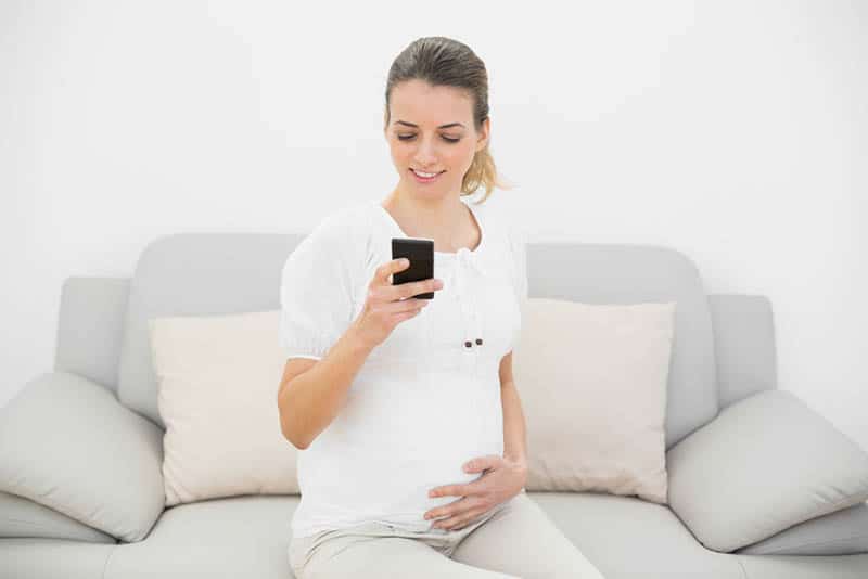 happy pregnant woman sitting on sofa and looking at mobile phone