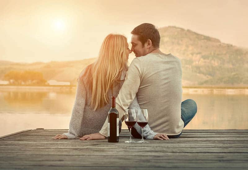romantic couple sitting by the lake with glasses and bottle of wine