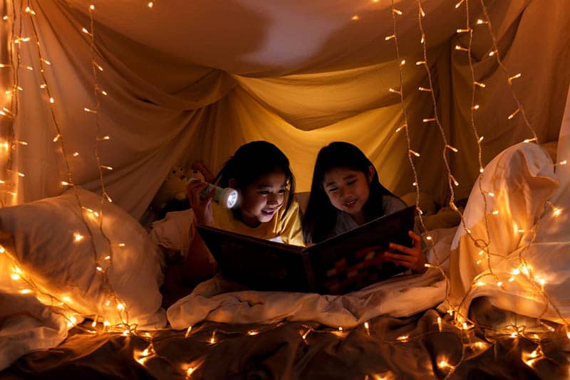 two sisters reading a book in a playing tent with flashlight