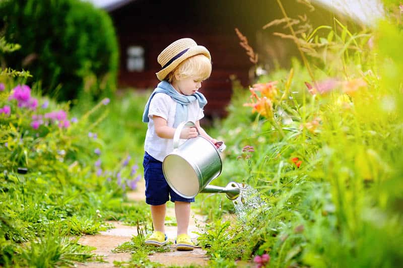 Cute toddler boy in straw hat watering plants in the garden