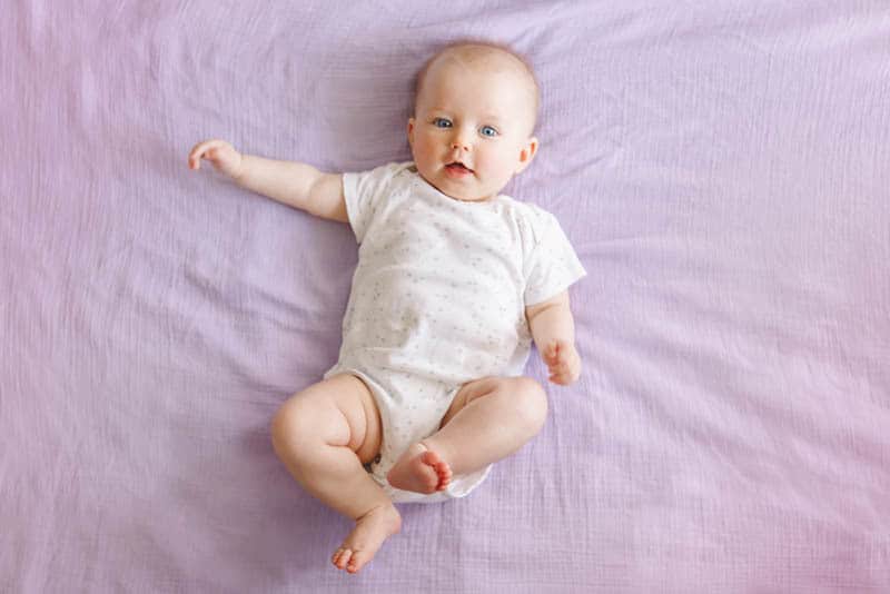 cute baby wearing white onesie and lying on purple sheet