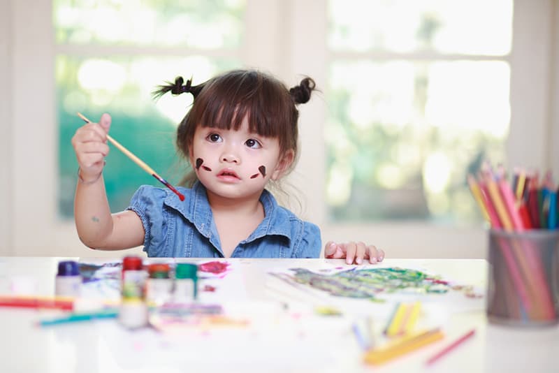 cute little girl holding a paint brush and sitting by the table