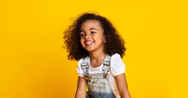 little girl with curly brown hair smiling in front of a yellow background
