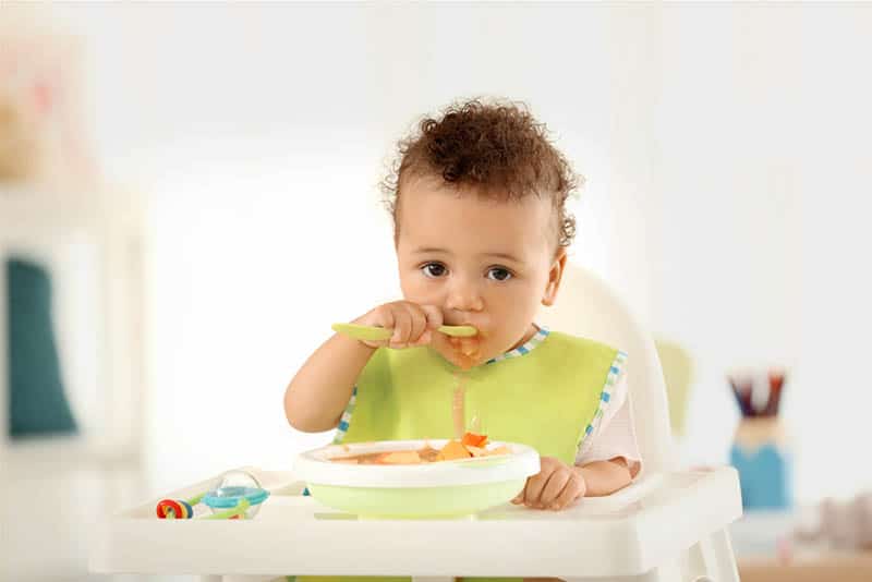 cute baby boy eating alone with spoon in high chair at home
