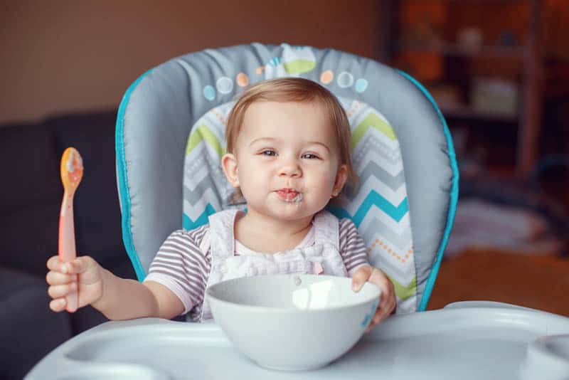 cute baby girl sitting in a high chair with bowl food in front and spoon in hand