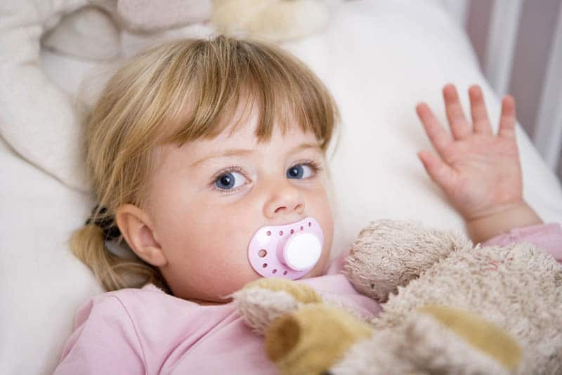 blond little girl with pacifier lying in bed with toy covered with blanket