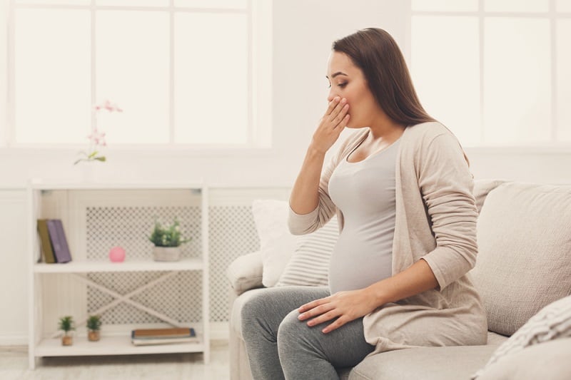 sick pregnant woman sitting on the couch and covering her mouth